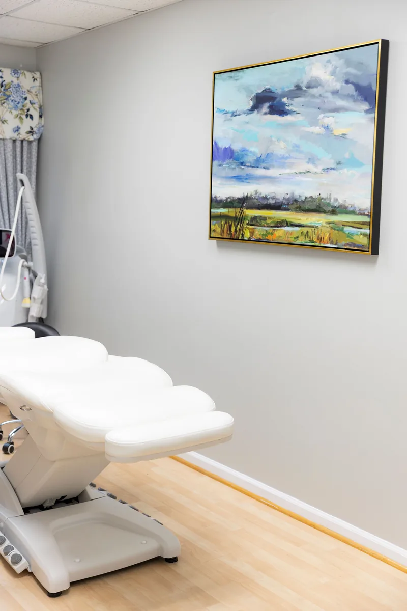 A treatment room at Colonial Aesthetics with a white treatment bed and a framed landscape painting