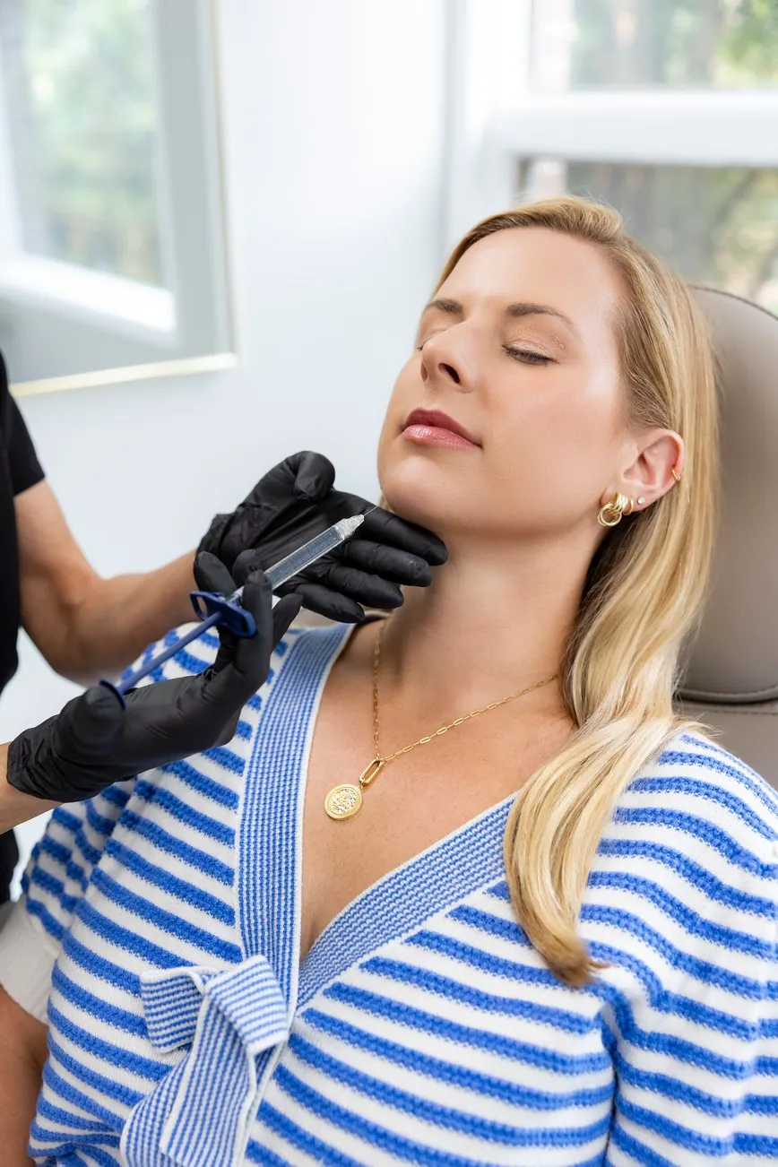 A patient reclines with her eyes closed while a gloved injector places a syringe along her jawline